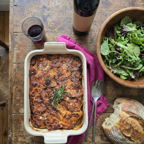 moussaka d'aubergines in a white ceramic casserole dish beside a tossed salad in a wooden dish and a crusty loaf of bread